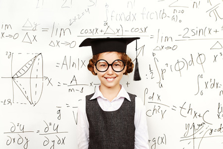 Happy smart schoolgirl in big glasses and academic hat performs the task at the blackboard. Education.の写真素材