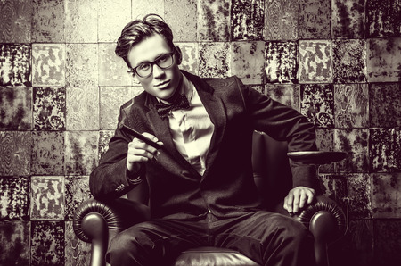 Handsome young man in elegant suit smoking a cigar. He is sitting on a leather chair in a luxurious interior.の写真素材