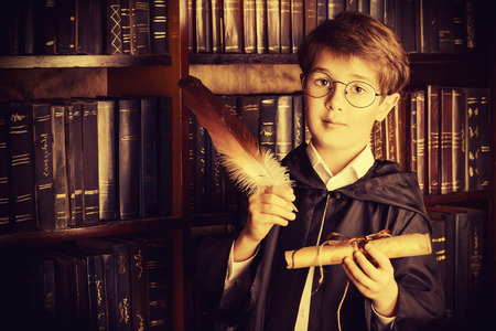 A boy stands in the library by the bookshelves with many old books and holds old manuscriptsの写真素材
