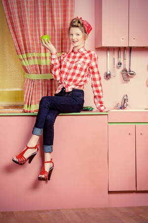 Pretty girl teenager wearing clothes and hair in pin-up style posing on a pink kitchen. Beauty, youth fashion. Pin-up style.の写真素材