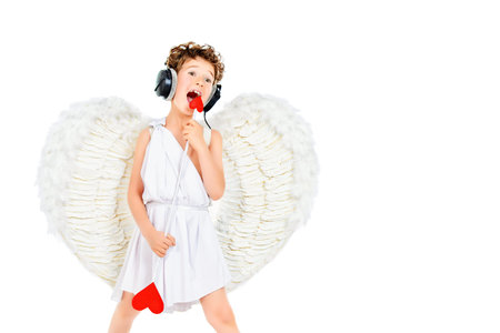 Cheerful little boy in a costume of angel listening to music in headphones and singing. Cupid....