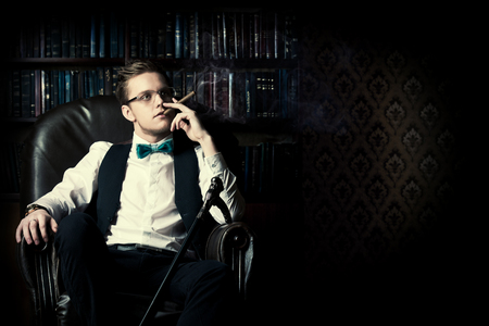 Elegant young man in a suit sitting in armchair and smoking a cigar. Vintage room. Fashion.の写真素材