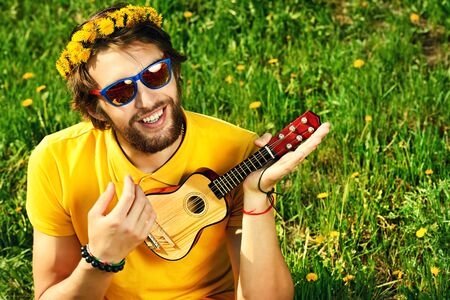 Happy carefree young man sitting on a grass and playing his little guitar. Summer, freedom, hippie.の写真素材