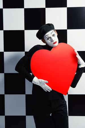 Portrait of a male mime artist holds large red heart expressing love. Chess board background.の写真素材