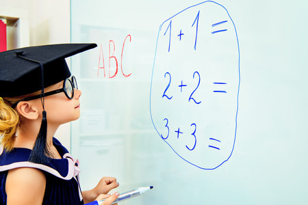 Cute little student girl in academic hat during the lesson at school. Educational concept.