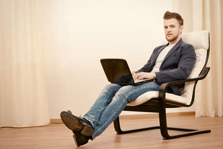 Happy young man using laptop sitting on armchair at home.の写真素材
