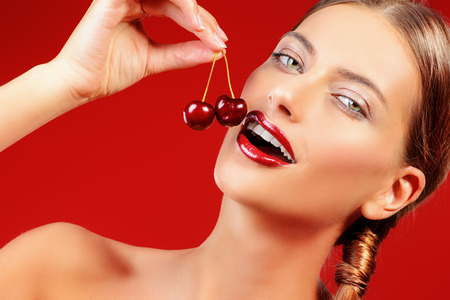 Attractive young woman eating fresh cherry. Sexual lips, red lipstick. Healthy food concept. Cosmetics. Red background.の写真素材