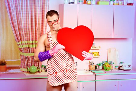 Handsome muscular man in an apron holding big red heart. Pink kitchen. Love concept. Valentine's day.の写真素材