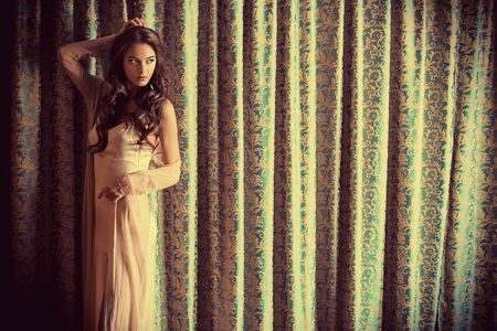 Attractive brunette woman in gorgeous evening dress posing by the vintage hangings. Beauty, fashion.の写真素材