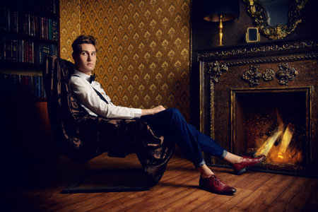 Elegant handsome young man sitting by the fireplace in a room with classic vintage interior