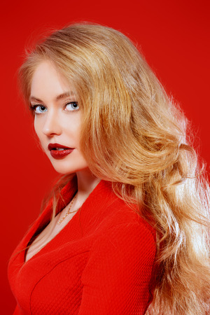 Beautiful young woman in red dress and with blonde curled hair. Beauty, fashion. Cosmetics, make-up. Red background.の写真素材
