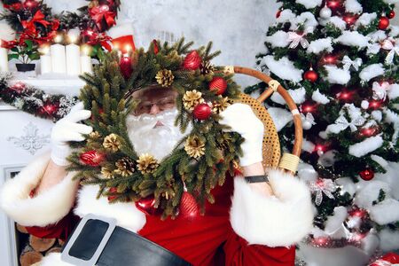 Happy Santa Claus sitting in a rocking chair with Christmas wreath in his arms. He is in the room by the fireplace and Christmas tree, beautifully decorated for Christmas.の写真素材
