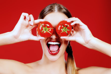 Beautiful laughing woman holding two ripe tomatoes before her eyes. Red background.の写真素材