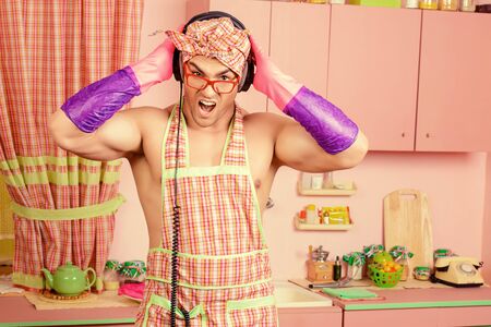 Funny and handsome muscular man in an apron takes a break from household chores, listening to music on headphones.の写真素材