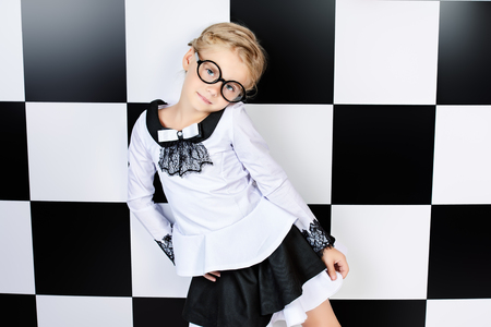 Portrait of a pretty little girl in white shirt and black skirt posing over chessboard background.の写真素材