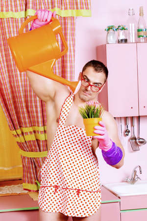 Muscular man in an apron watering the flowers with expression. He is in the pink kitchen.の写真素材