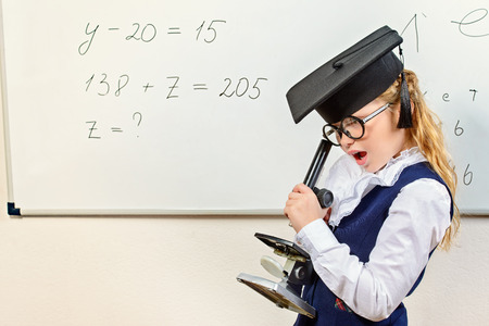 Cute smart schoolgirl in big glasses and academic hat stands at the blackboard and curiously looking into the microscope. Education.の写真素材