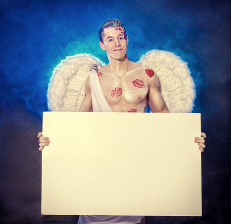 Portrait of a male Angel Cupid holding white billboard and smiling at camera. Valentine's day. Love...