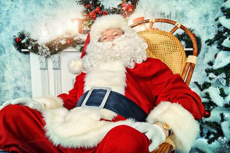 Good old Santa Claus sitting in a rocking chair in the room by the fireplace and Christmas tree, beautifully decorated for Christmas.の写真素材