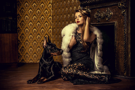 Beautiful woman in the precious jewelry and fur coat sitting by the fireplace with her dog. Classic...