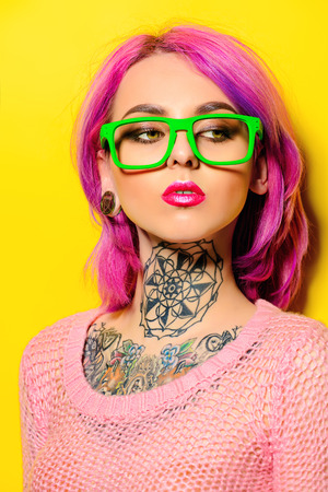 Attractive young woman with crimson hair wearing bright green glasses posing over yellow background. Bright style, fashion. Optics style. Tattoo. Hair coloring.の写真素材
