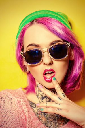 Bright fashion concept. Trendy girl with crimson hair wearing bright clothes and sunglasses alluring by a  yellow background. Optics style. Tattoo.の写真素材