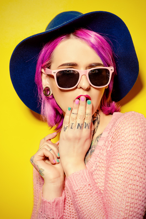 Pretty girl with crimson hair wearing bright clothes and sunglasses posing over yellow background. Bright style, fashion. Optics style. Tattoo.の写真素材