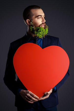 Elegant young man with a beard of green flowers holding red heart. Love concept. Valentine's Day. Barbershop.の写真素材