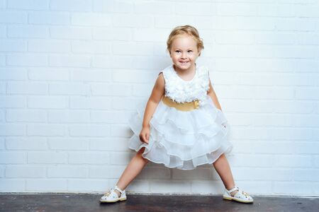 Angelic little girl in a beautiful white dress standing by the white brick wall. Childhood.の写真素材