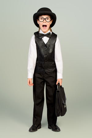Portrait of the old styled boy in elegant suit and bowler hat with his suitcase. Studio shot. Kid's fashion.の写真素材