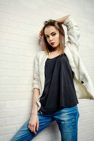 Modern young woman in casual jeans and jacket posing by a white brick wall.の写真素材