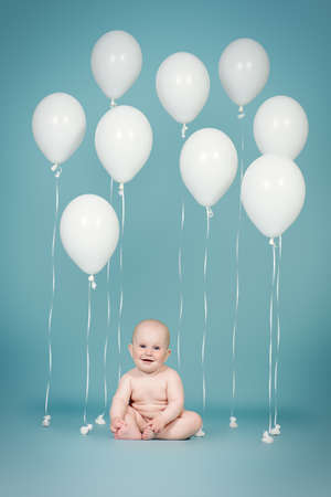 Adorable little baby sitting among white balloons over blue background. Infant baby. Healthcare. Happy childhood.の写真素材