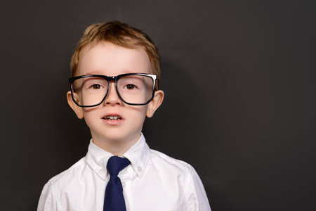 Four year old boy in white shirt and a tie posing by a school blackboard in big glasses. Educational...