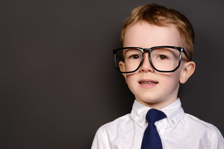 Four year old boy in white shirt and a tie posing by a school blackboard in big glasses. Educational...