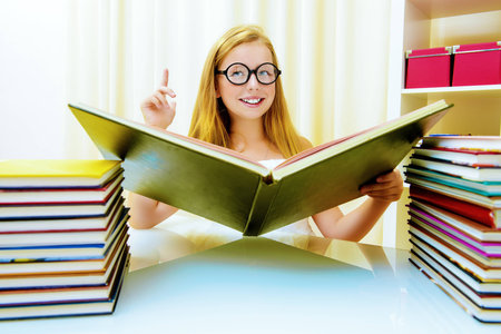 Teen girl sitting at table at home with piles of books and smiles. Educational concept.