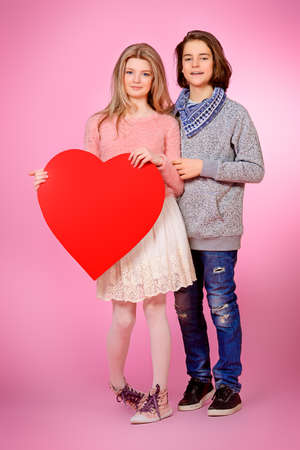 Full length portrait of happy teenage boy and girl holding red heart over pink background. Friendship. First love. Valentine's Day.の写真素材