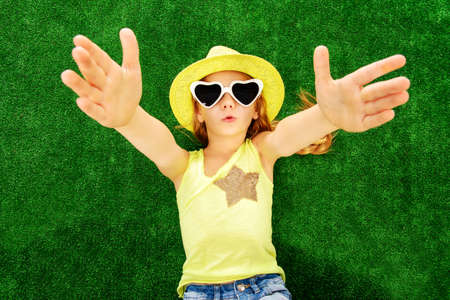 Joyful little girl in colorful clothes and sunglasses lying on a green lawn. Kid's fashion. Summer holidays.の写真素材