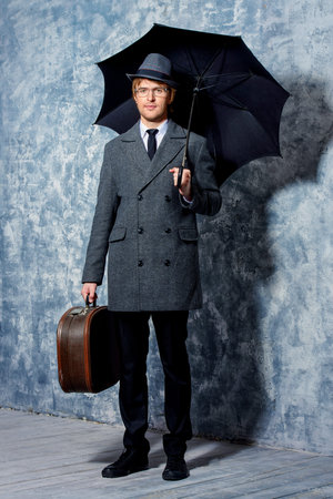 Elegant young man wearing classic hat and a coat stands with his black umbrella. Beauty, fashion.の写真素材