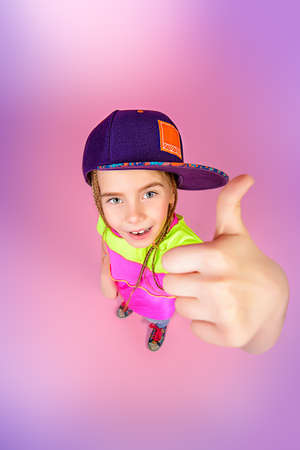 Cute little girl in a cap smiling to camera and showing thumbs. Kid's style. Happy childhood.の写真素材