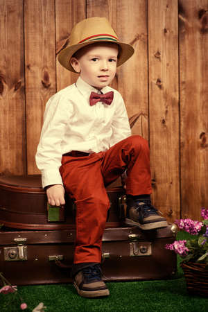 Cute little boy in elegant clothes sitting on his old suitcases on a green lawn with flowers over wooden background. Kid's fashion. Childhood. Summer holidays.の写真素材