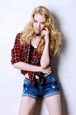 Sexual blonde girl in jeans shorts and checkered shirt alluring by a brick wall. Beauty, fashion.の写真素材