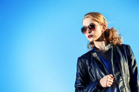Modern young woman in leather jacket and sunglasses posing outdoor over blue sky. Beauty, fashion. Urban style.の写真素材