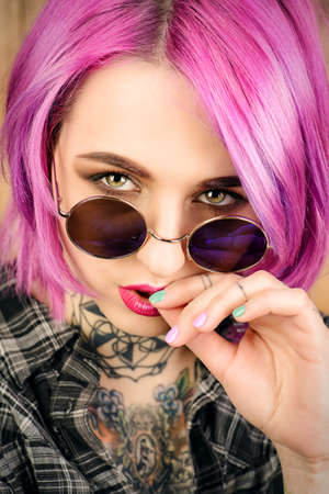 Close-up portrait of a modern girl with bright crimson hair and round sunglasses. Tattoo. Hair coloring. Optics style.の写真素材