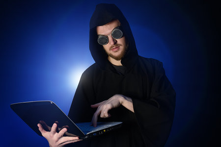 A hacker hiding his face behind glasses and a hood typing on his laptop. Programmer.