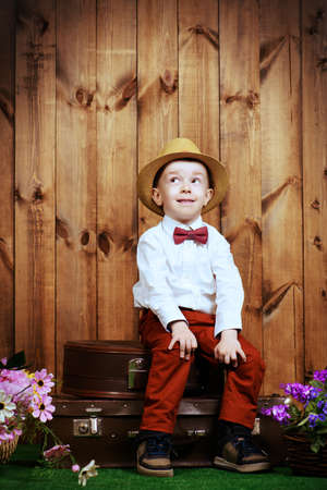 Cute little boy in elegant clothes sitting on his old suitcases on a green lawn with flowers over wooden background. Kid's fashion. Childhood. Summer holidays.の写真素材