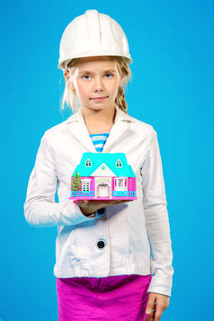 Little girl in a builder helmet  holding house in her hands. The construction industry, mortgage concept. The house for the whole family.の写真素材