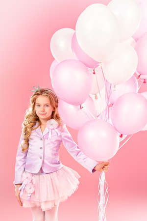 Pretty little girl with beautiful blonde hair alluring with pink balloons over pink background. Little princess with a crown on her head. Kids fashion. の写真素材