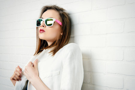 Modern brunette young woman in sunglasses posing by a white brick wall. Beauty, fashion. Youth...
