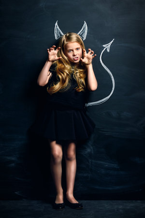 Funny little imp girl. Cute child girl posing with imp horns and tail drawn on a blackboard.