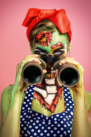 Portrait of a pin-up zombie woman looking through binoculars, pink background. Body-painting project. Glamorous zombie girl. Halloween make-up.の写真素材